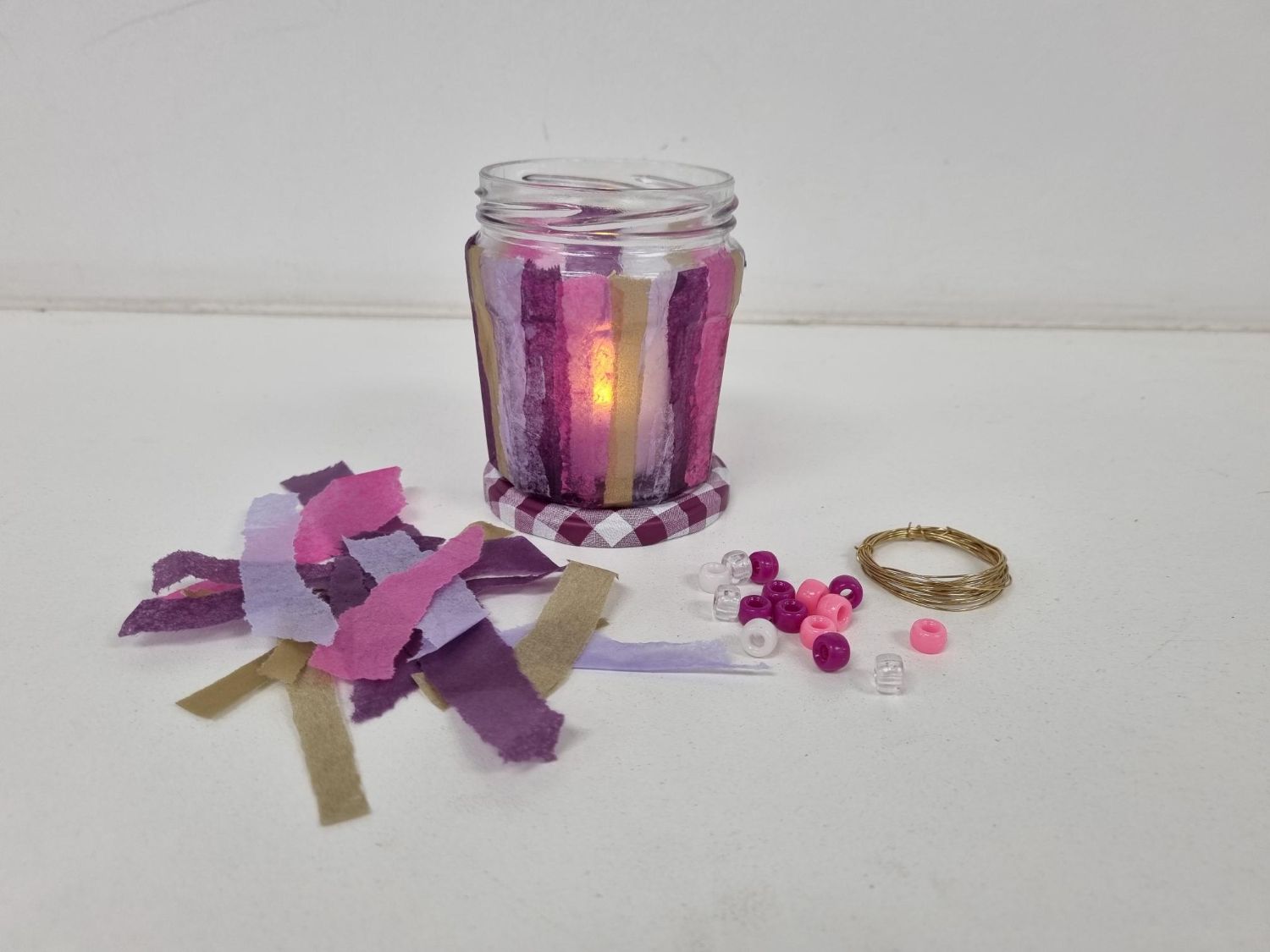 A glass jar candle holder is decorated with vertical strips of purple, pink, and gold tissue paper, illuminated by a glowing tealight inside.